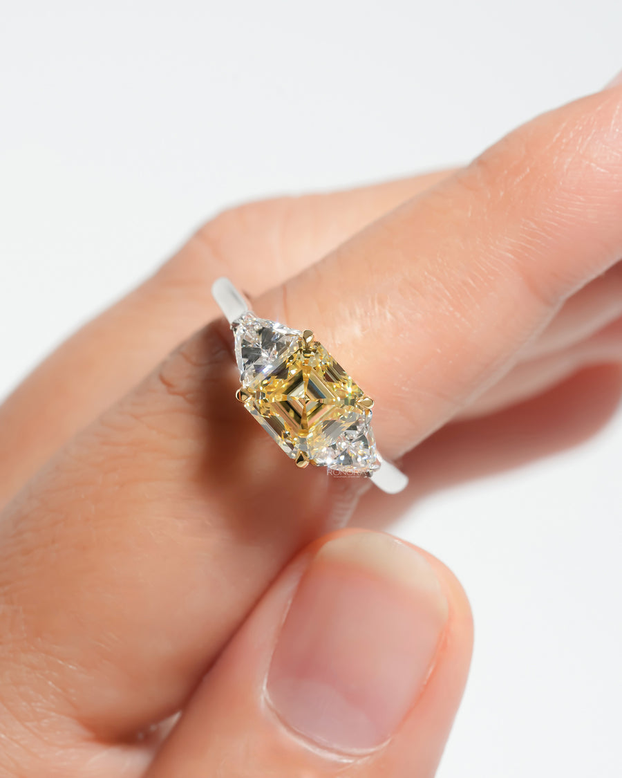 Fancy yellow asscher cut lab grown diamond ring with two white side stones, held between fingers, close-up