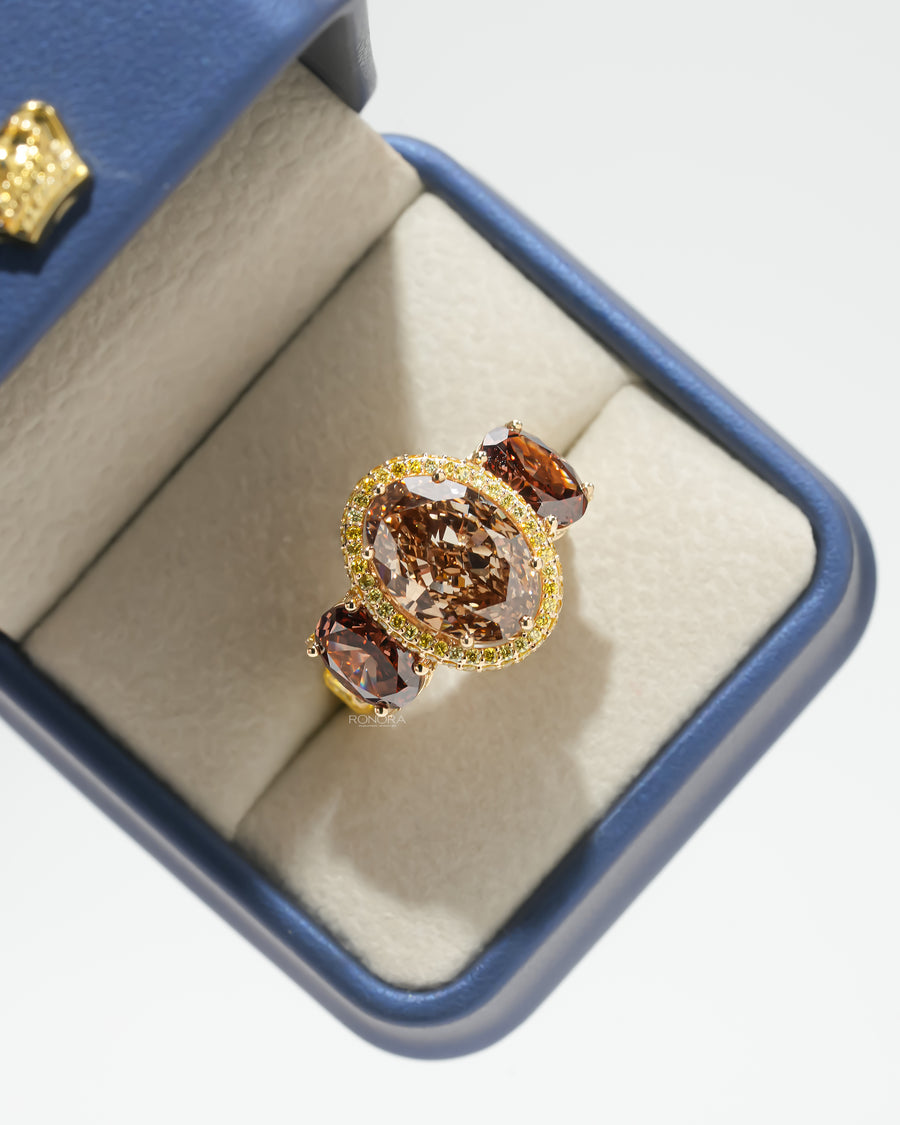 Brown oval lab grown diamond three stone ring with yellow halo in a navy blue ring box, seen from above