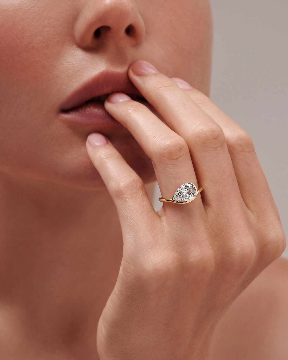 Model wearing an east west pear lab grown diamond ring in yellow gold, fingertips resting on her lips