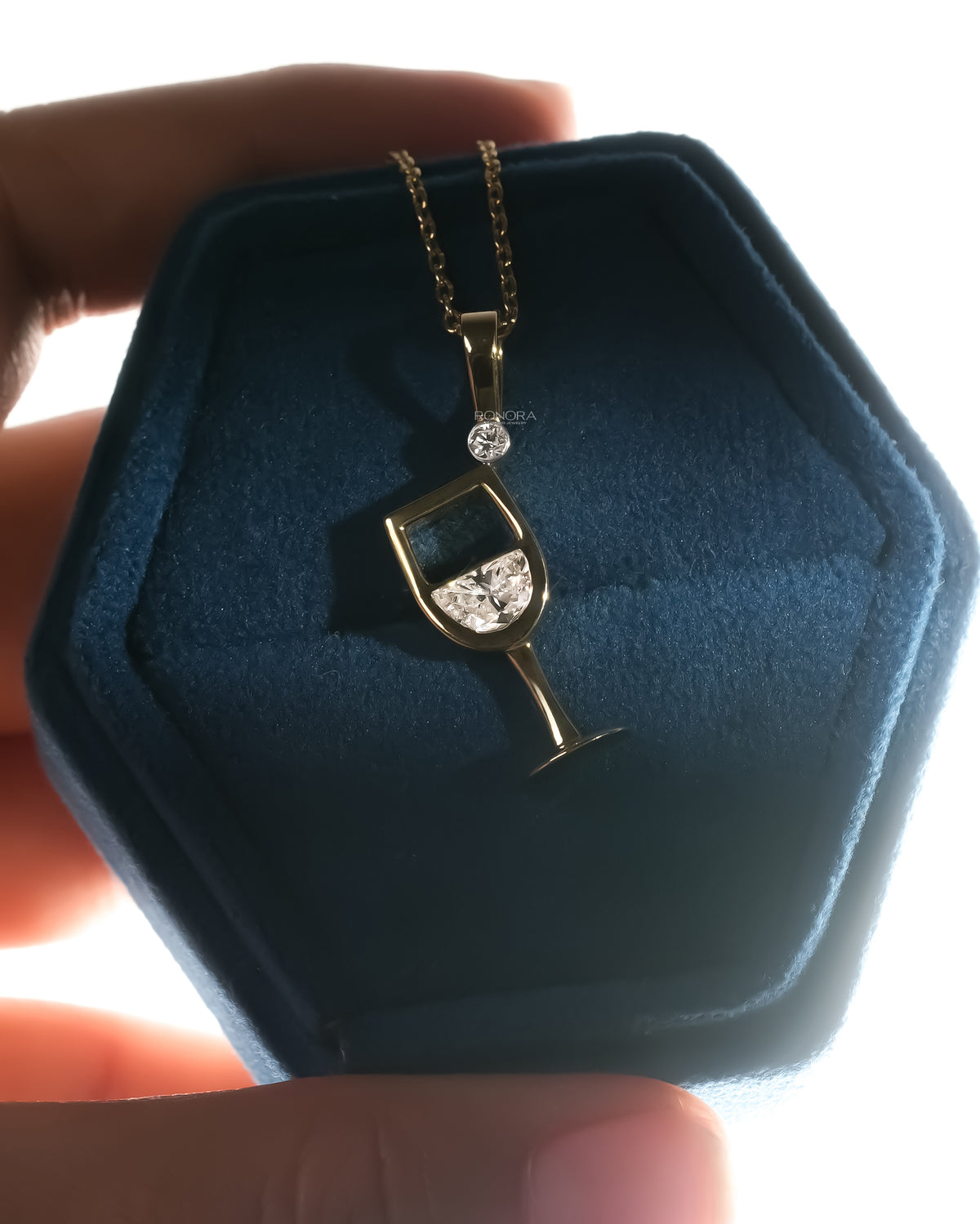 Hand holding a dark blue velvet box with a yellow gold wine glass lab grown diamond pendant, backlit