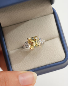 Asscher yellow lab grown diamond ring with white heart side stones in an open navy ring box held by a hand