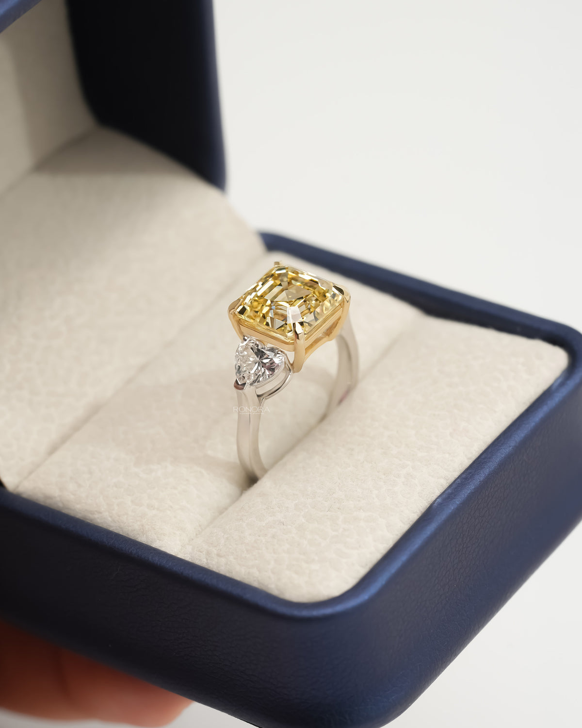 Asscher yellow lab grown diamond ring in an open navy ring box, yellow gold prongs and a white metal band