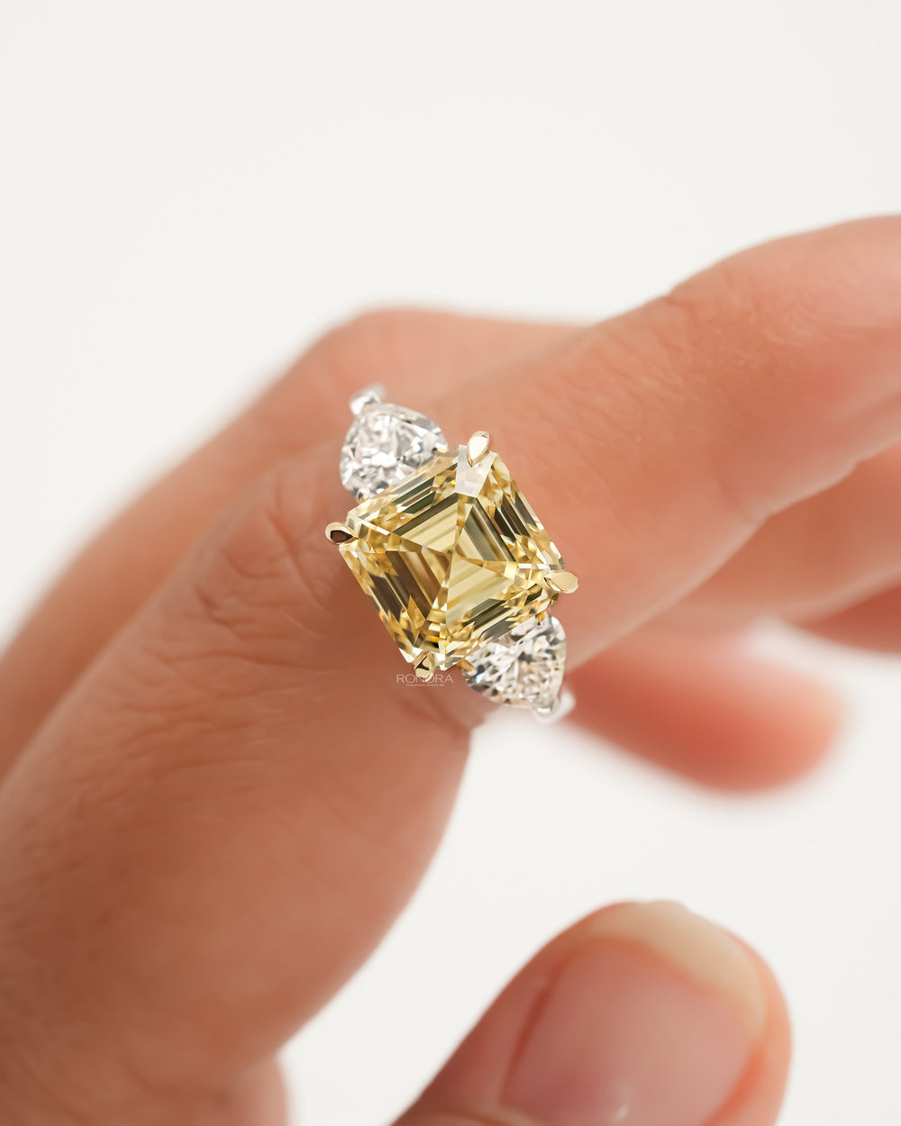 Close-up of an asscher yellow lab grown diamond ring on a finger, yellow gold prongs and white heart side stones