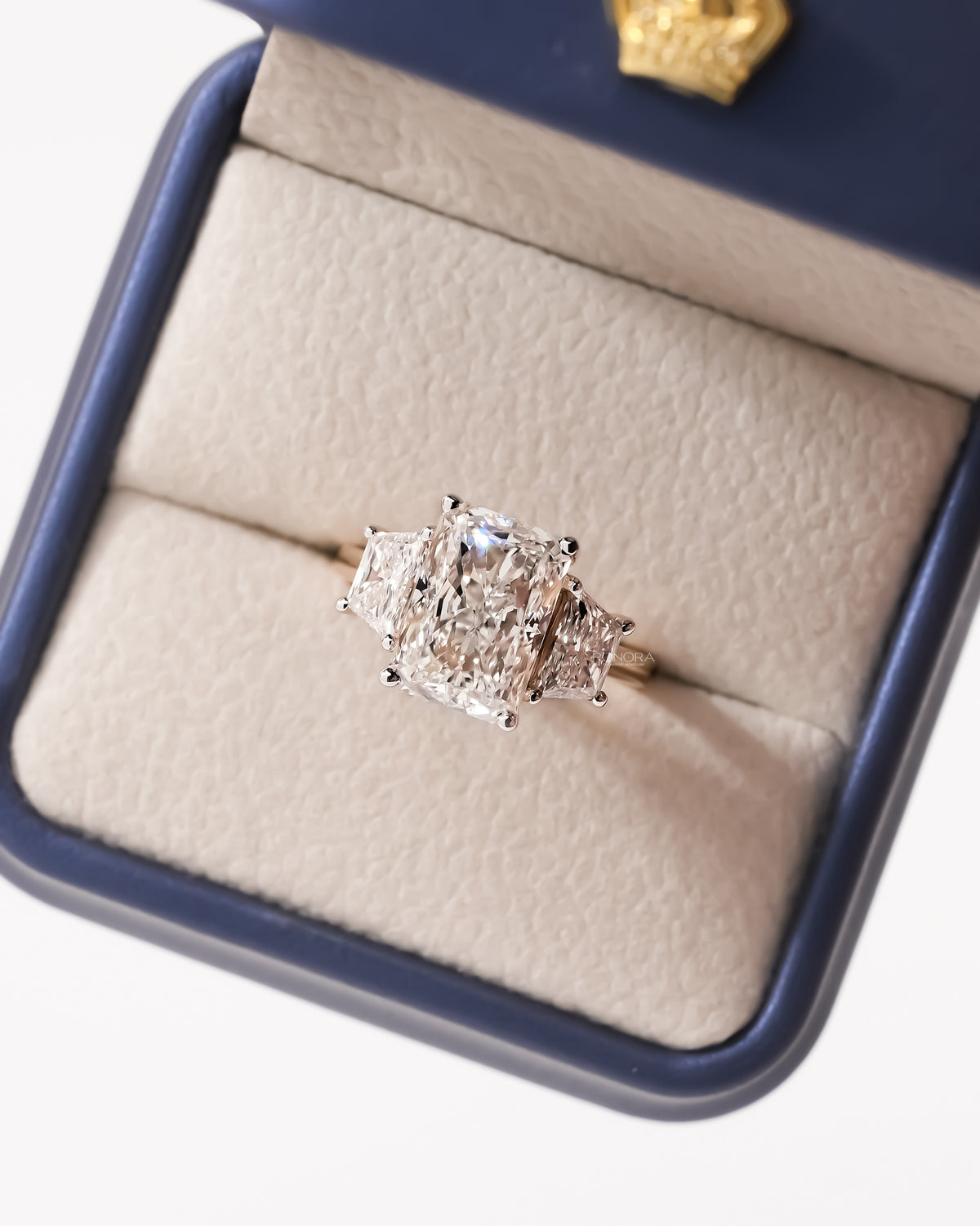 Top view of a radiant cut lab grown diamond three stone ring with trapezoid side stones in a navy ring box