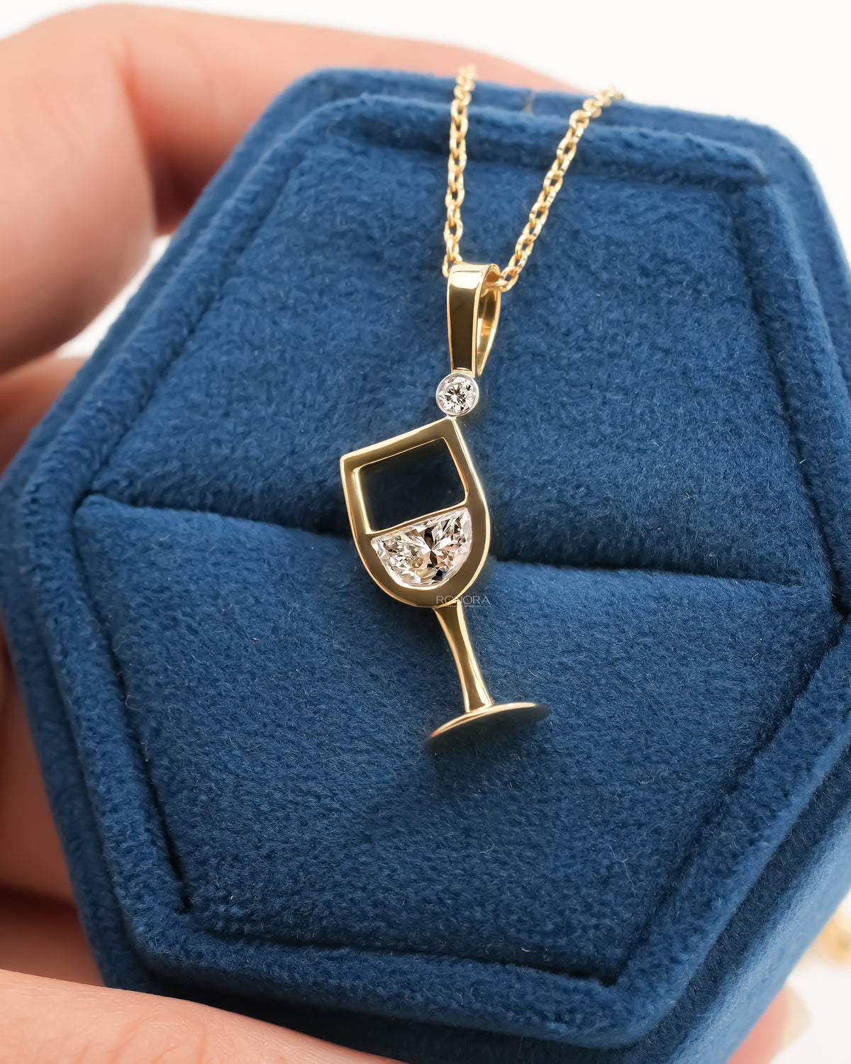 Wine glass lab grown diamond pendant in yellow gold on a blue velvet ring box held in a hand