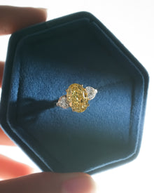 Oval yellow lab grown diamond ring with heart side stones in a blue velvet hexagon jewelry box held in fingers