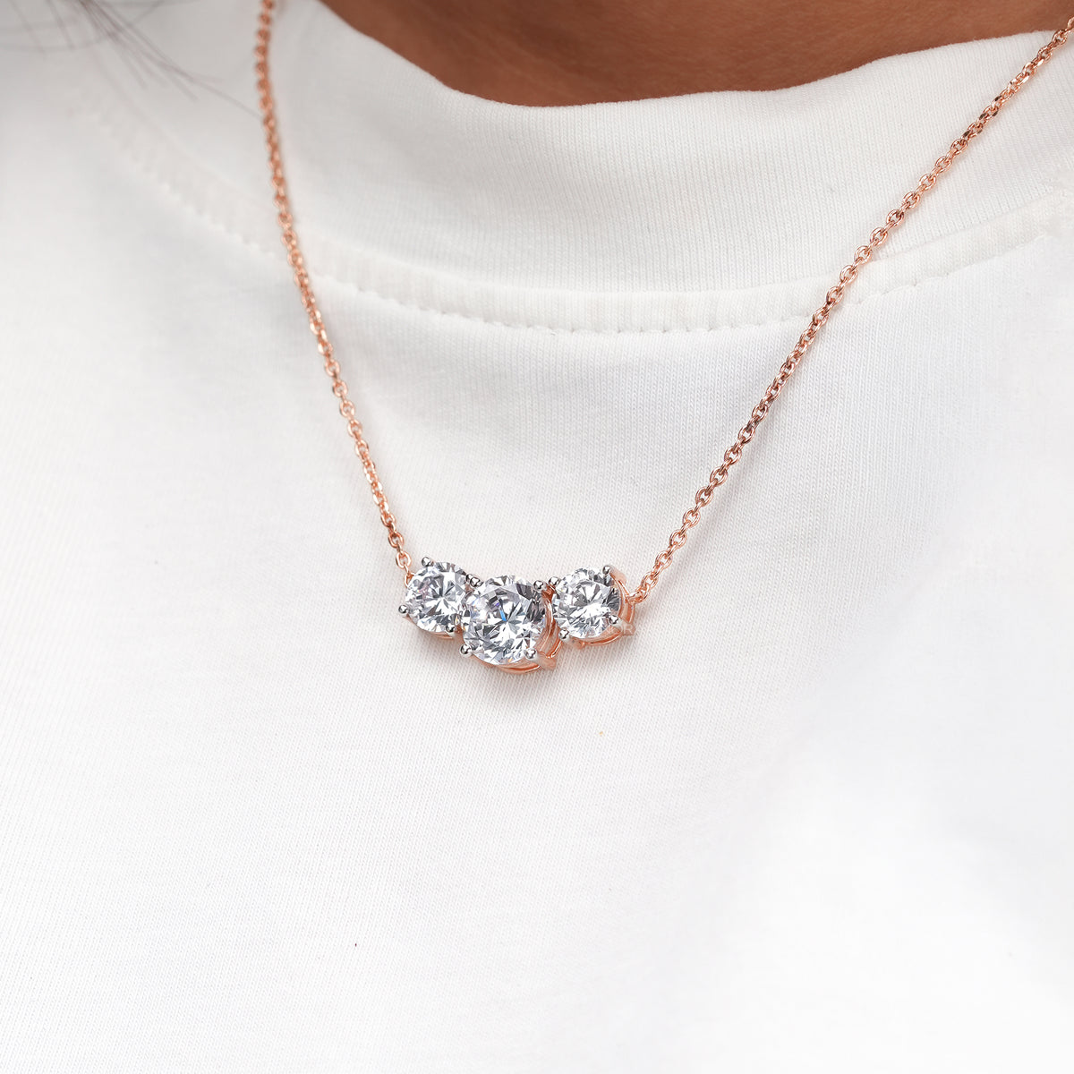Close-up of a rose gold three stone round lab grown diamond necklace worn on a model in a white t-shirt