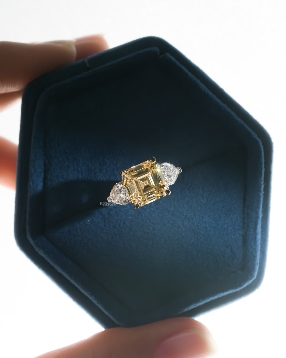 Asscher cut yellow lab grown diamond ring with white heart shaped side stones in a blue velvet hexagon jewelry box