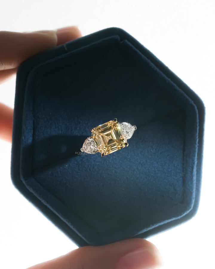 Asscher cut yellow lab grown diamond ring with white heart shaped side stones in a blue velvet hexagon jewelry box