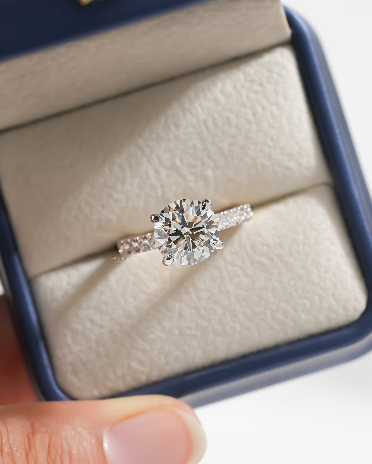 Top view of a round hidden halo lab grown diamond ring with pave band in a navy and cream ring box