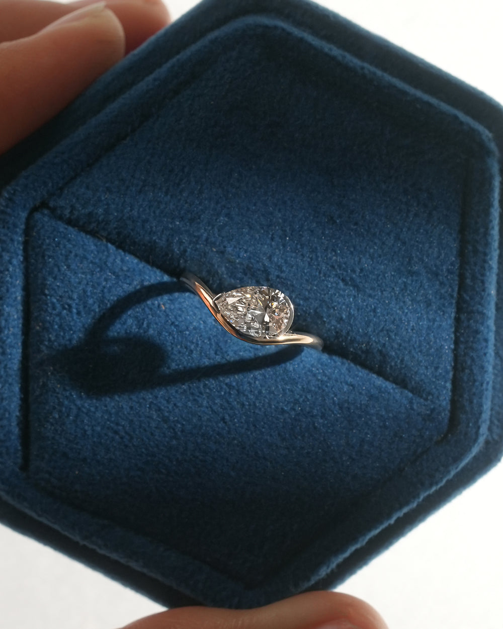 Top view of an east west pear lab grown diamond ring in a blue velvet hexagon jewelry box held in hand