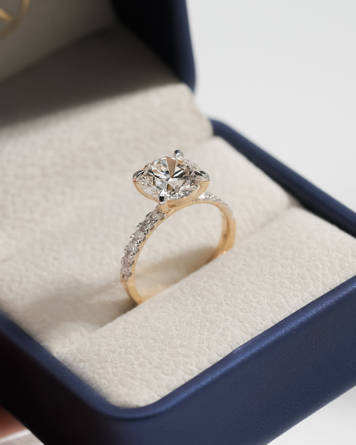 Round hidden halo lab grown diamond ring in yellow gold with a pave band, angled in a navy ring box
