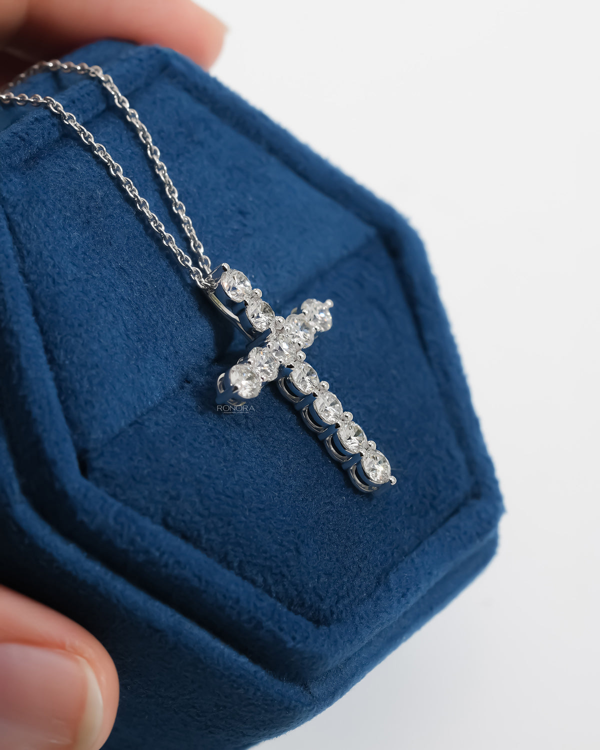 Side angle of a round lab grown diamond cross necklace in a blue velvet jewelry box showing the prong settings
