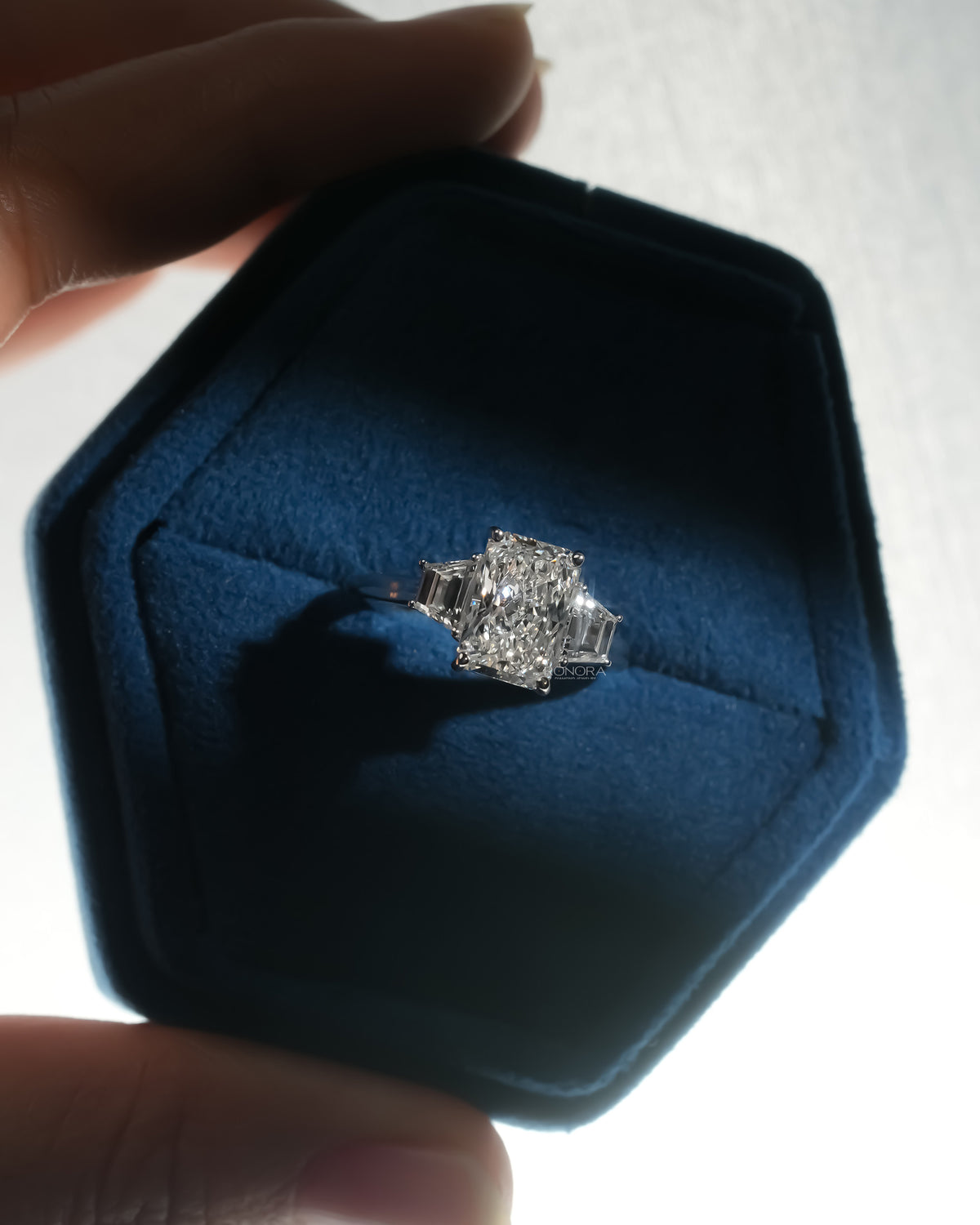 Radiant three stone lab grown diamond ring with trapezoid side stones in a blue velvet hexagon jewelry box