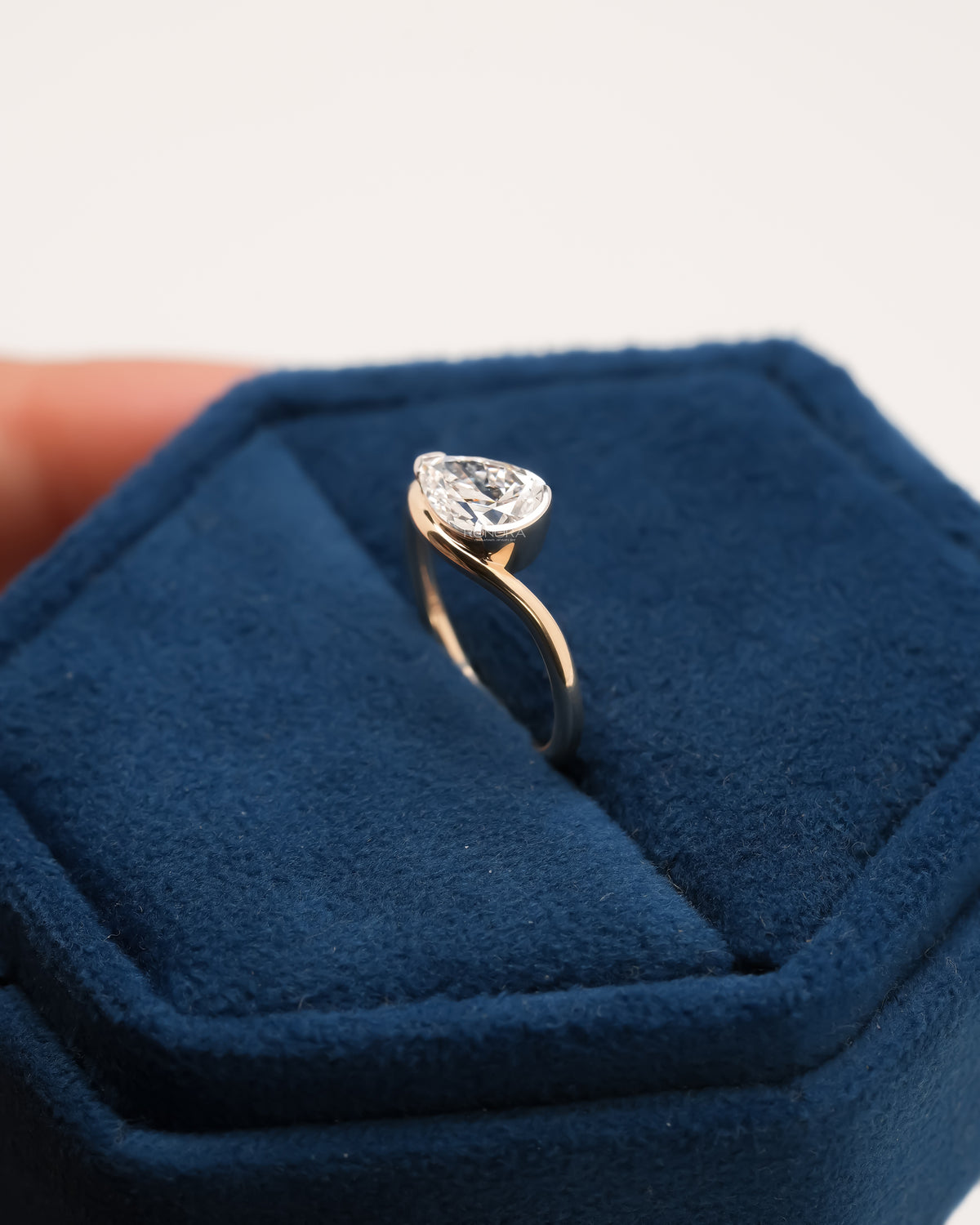East west pear lab grown diamond ring standing upright in a blue velvet jewelry box, gold band with a bezel stone