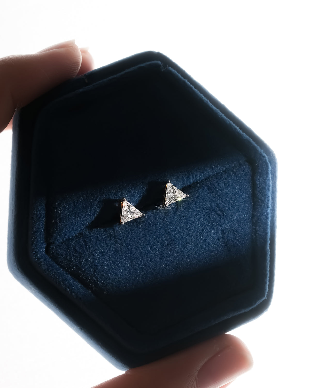 Trillion cut lab grown diamond earrings in a navy velvet hex box held between fingers, backlit