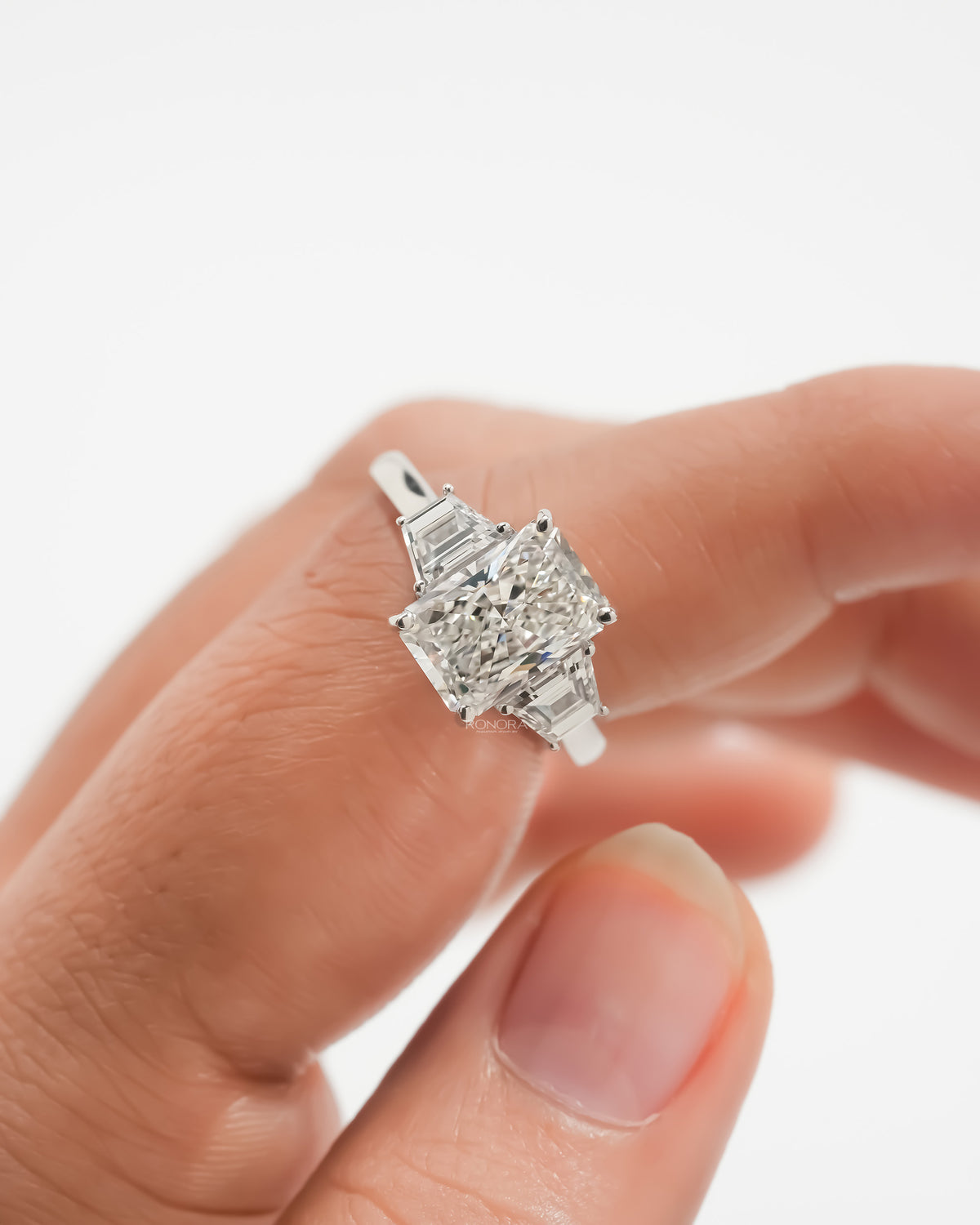 Close-up of a radiant three stone lab grown diamond ring with trapezoid side stones held on a finger