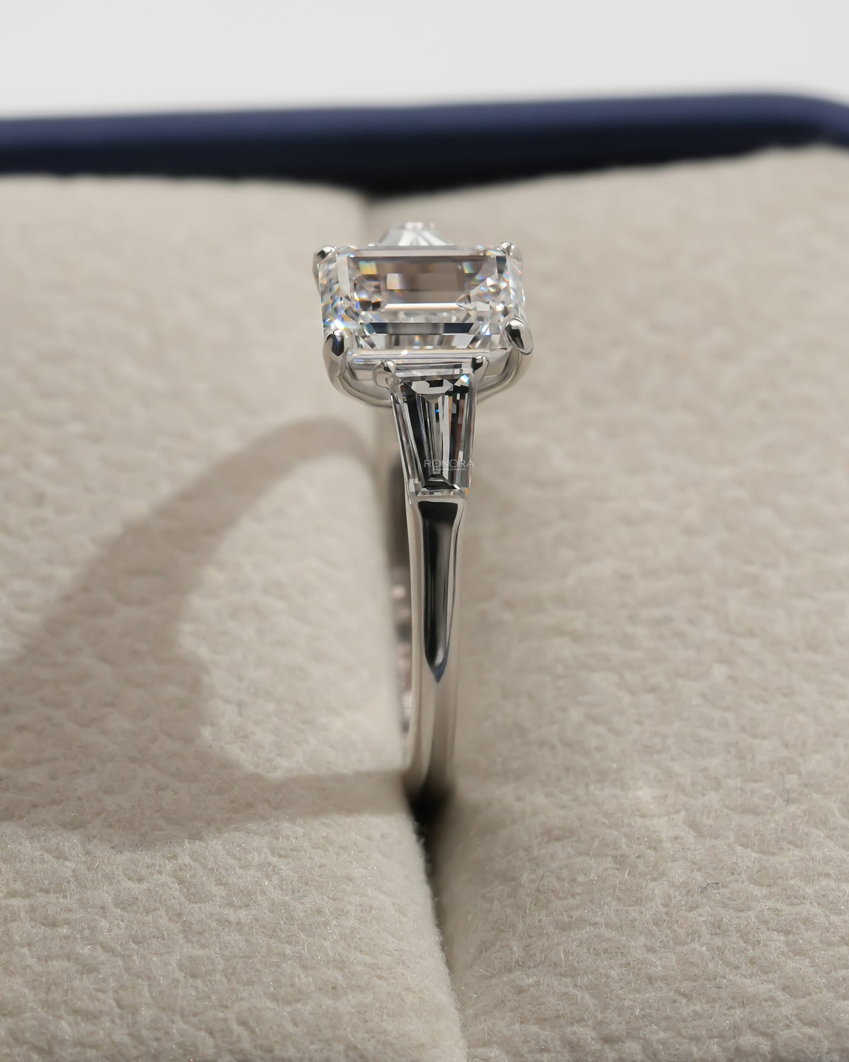 Side profile close-up of an emerald cut lab grown diamond three stone ring in a cream ring box with navy trim