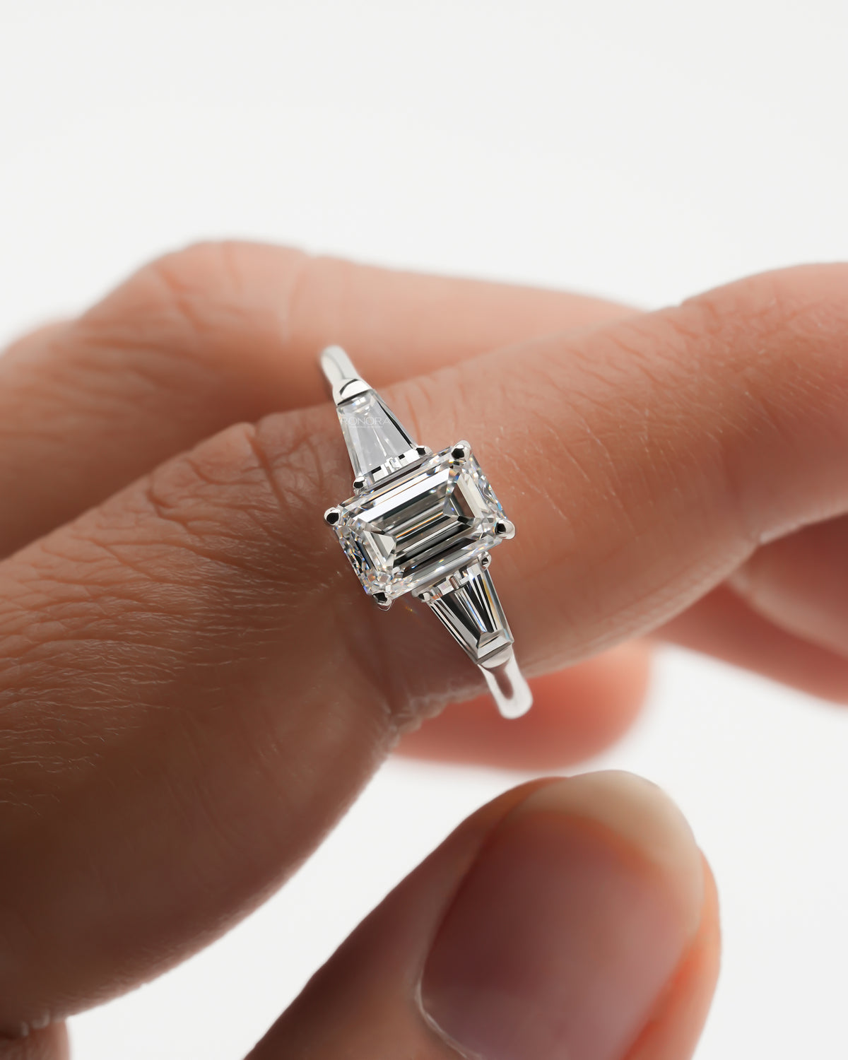 Emerald cut lab grown diamond three stone ring held between fingers, showing tapered baguette side stones