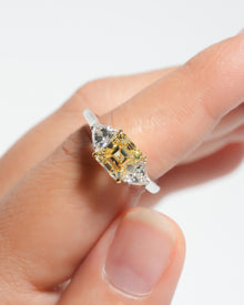 Fancy yellow asscher cut lab grown diamond ring with two white side stones, held between fingers, close-up