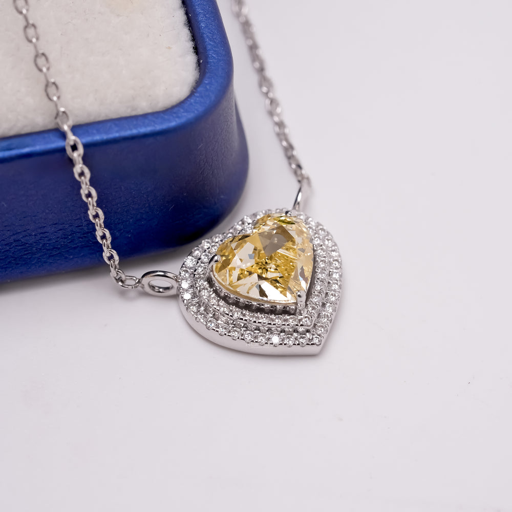 Close-up of a yellow heart cut lab grown diamond necklace with a double halo beside a blue jewelry box