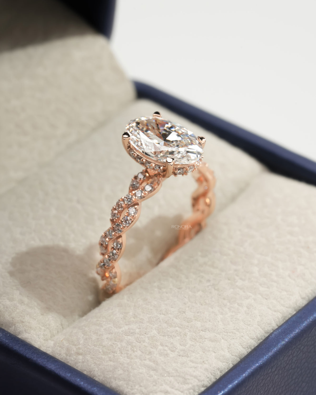 Angled view of an oval lab grown diamond ring with a rose gold twisted pave band, sitting in a navy and cream jewelry box