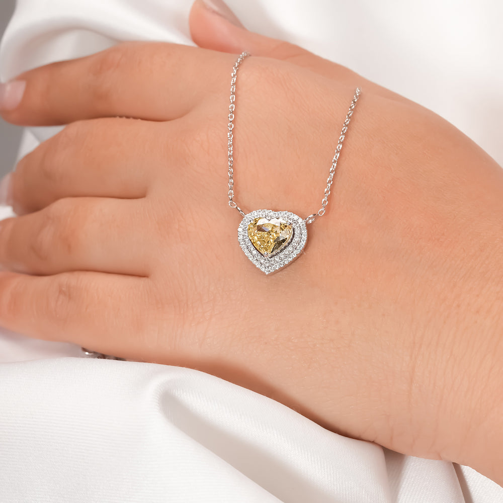 Yellow heart cut lab grown diamond necklace with a double halo worn on a hand resting on white satin fabric