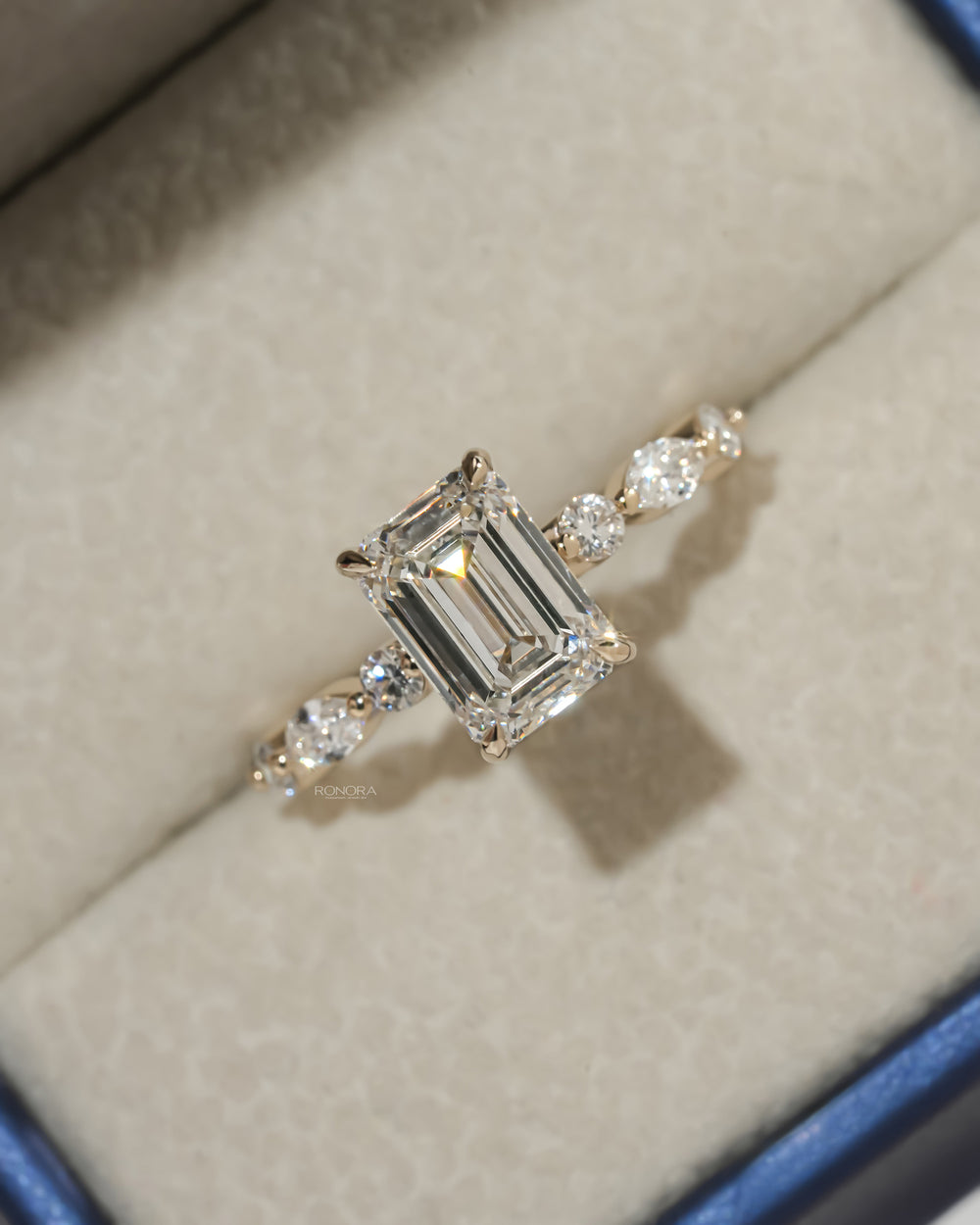 Close-up of an emerald cut lab grown diamond ring in yellow gold with side diamonds, resting in a cream jewelry box