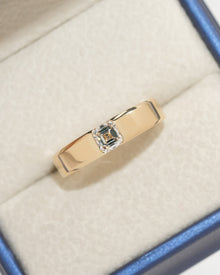 Top view of an asscher cut lab grown diamond ring in a wide polished yellow gold band, in a cream and blue jewelry box