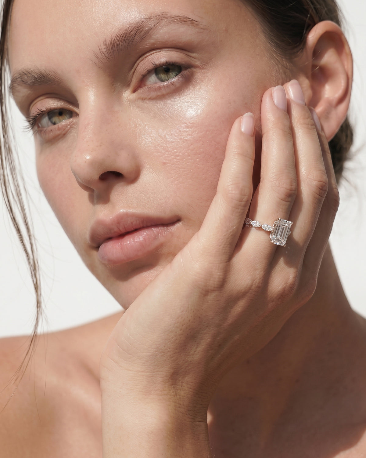 Woman resting her cheek on her hand, wearing an emerald cut lab grown diamond ring with side diamonds on a slim gold band