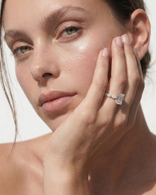 Woman resting her cheek on her hand, wearing an emerald cut lab grown diamond ring with side diamonds on a slim gold band