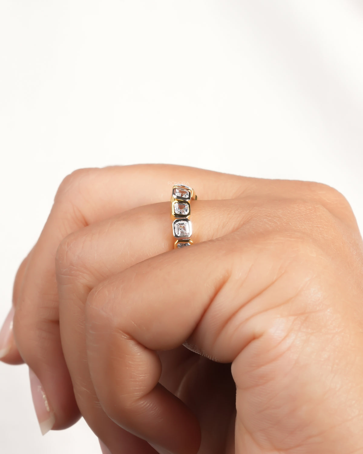 Angled view of an asscher cut lab grown diamond eternity band worn on a finger with bezel set square stones