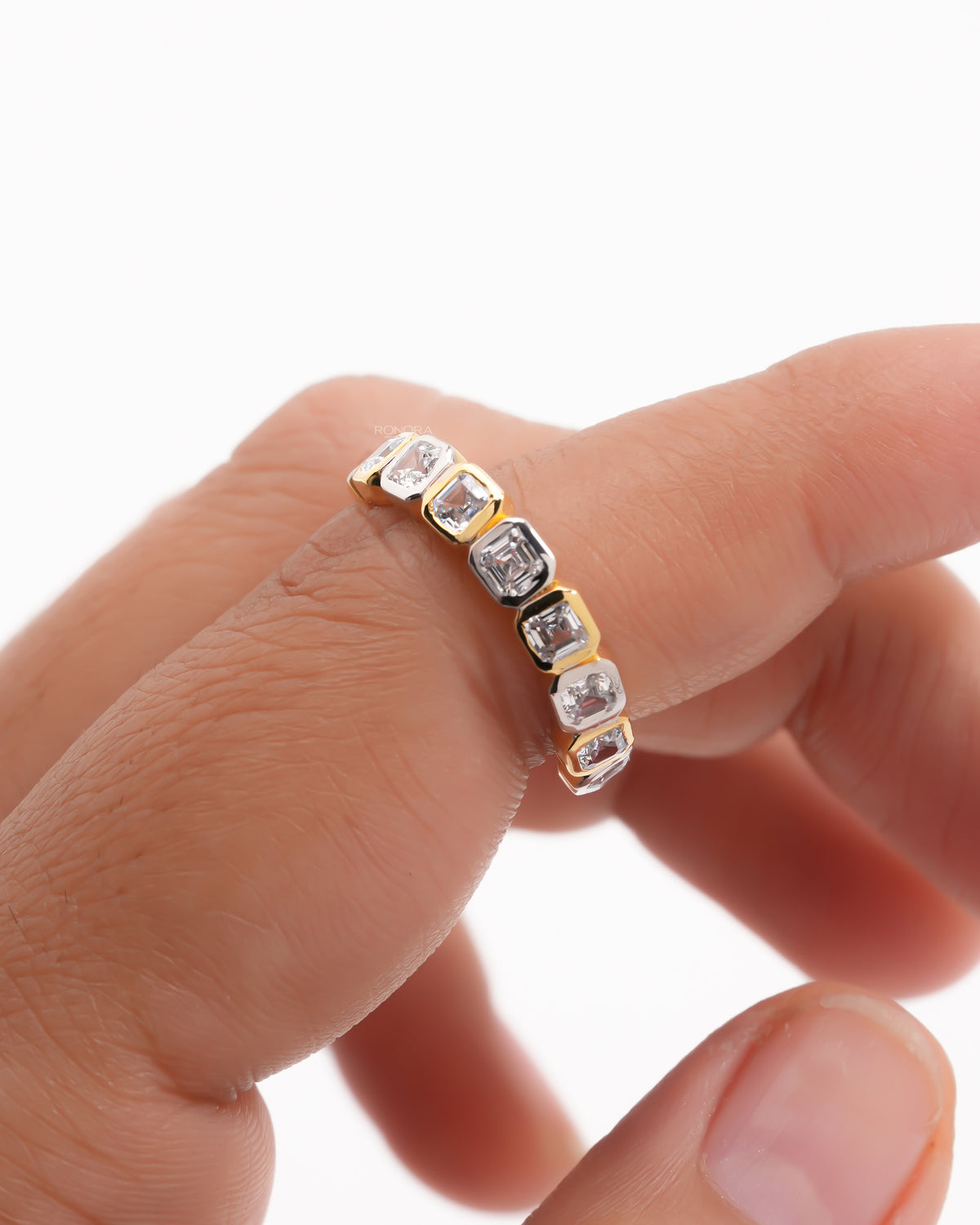 Close-up of an asscher cut lab grown diamond eternity band held on a fingertip with yellow and white gold bezels