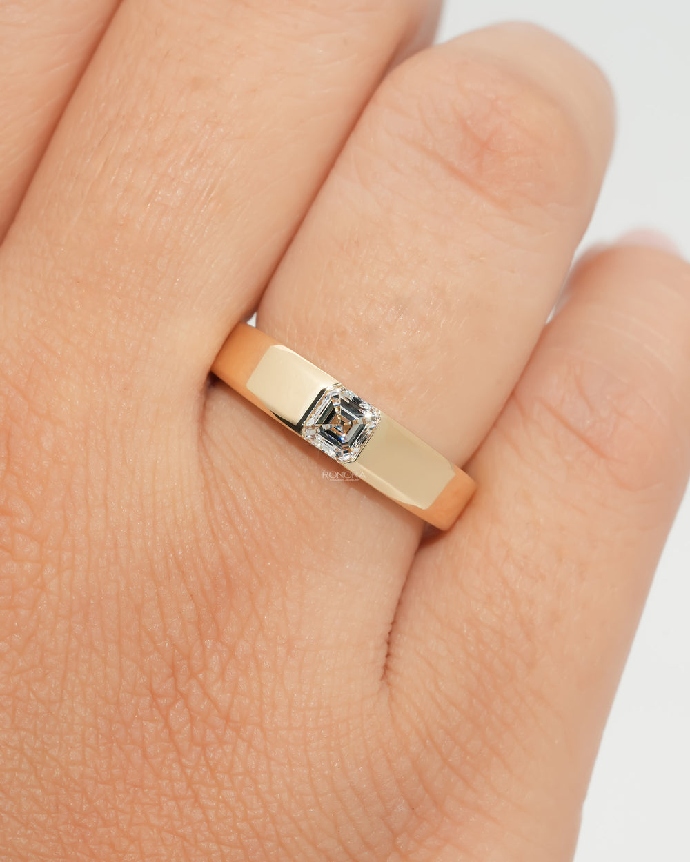 Close-up of an asscher cut lab grown diamond ring in a wide polished yellow gold band, worn on a hand