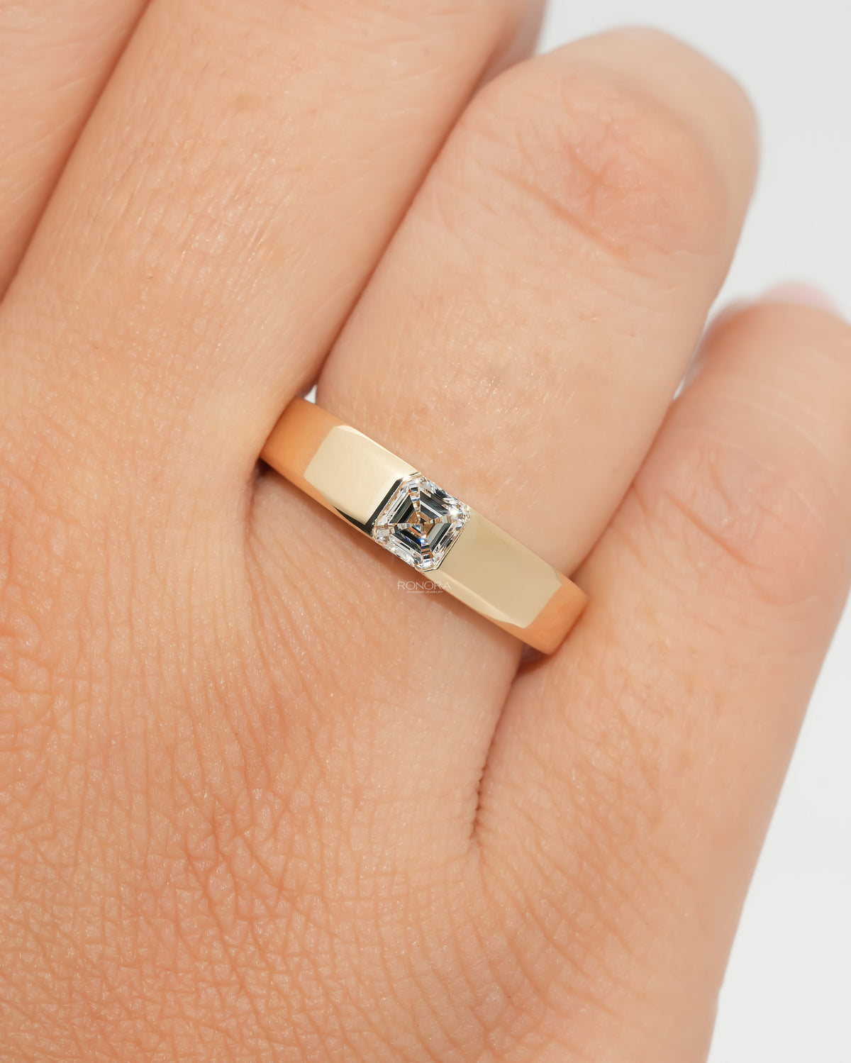 Close-up of an asscher cut lab grown diamond ring in a wide polished yellow gold band, worn on a hand