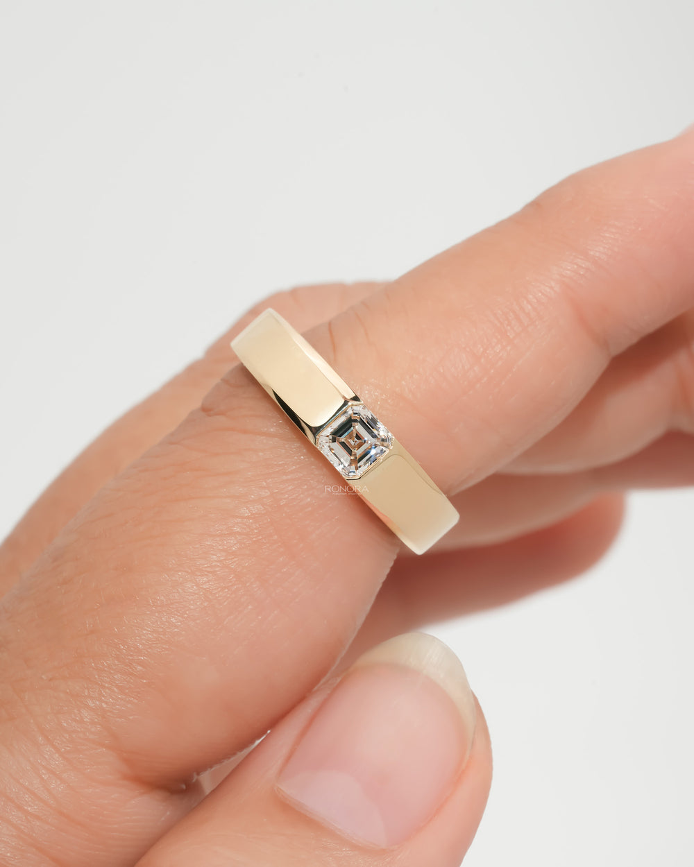 Asscher cut lab grown diamond ring in a wide yellow gold band, held between fingertips against a soft grey background