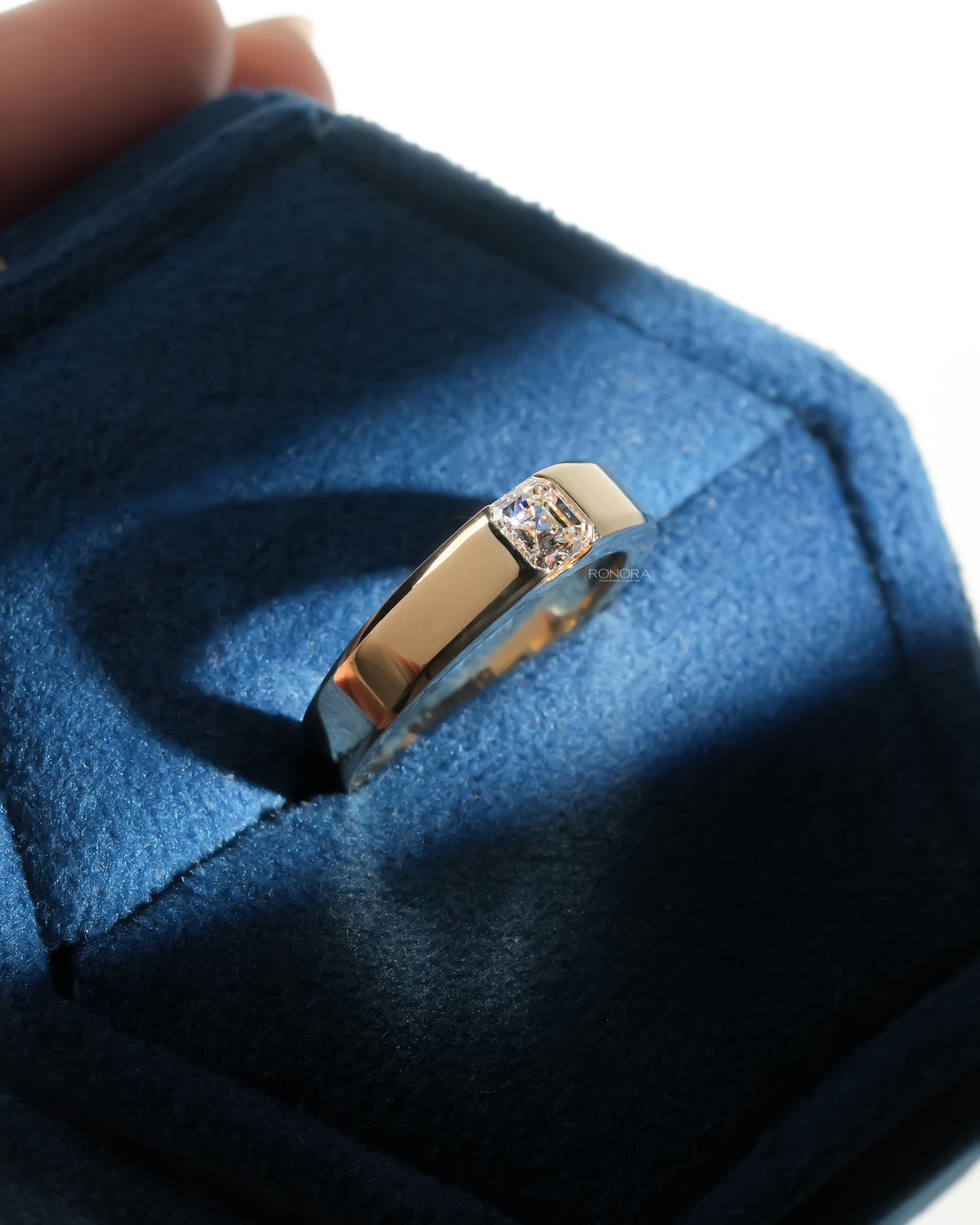 Angled view of an asscher cut lab grown diamond ring in a wide yellow gold band, resting in a blue velvet ring box