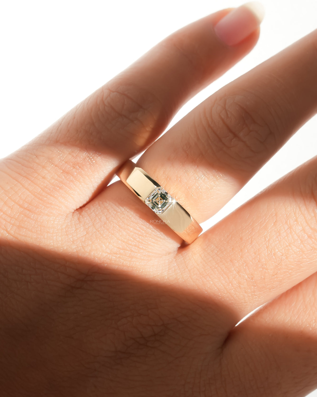 Asscher cut lab grown diamond ring with a bezel set stone in a wide yellow gold band, worn on a hand in warm sunlight