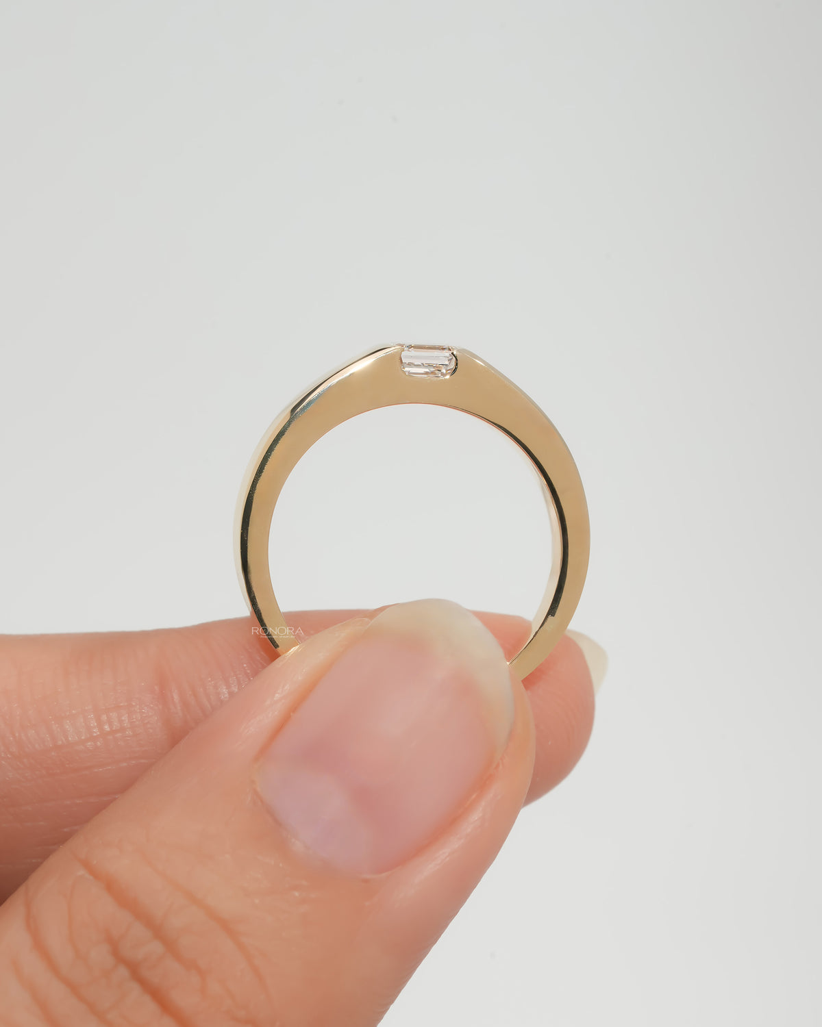 Asscher cut lab grown diamond ring held up in fingers, showing the full yellow gold band with the bezel set stone on top