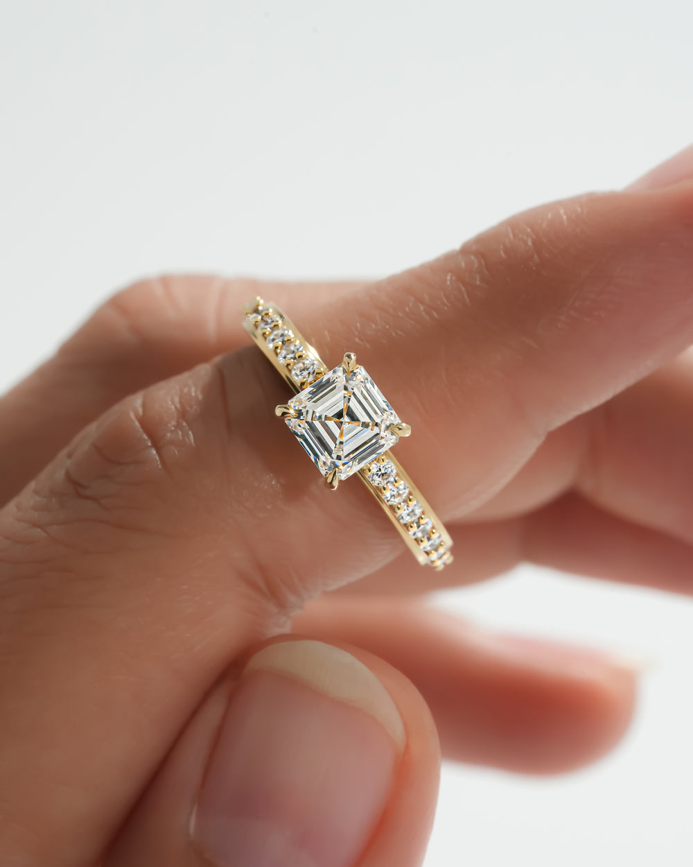 Close-up of an asscher cut pave lab grown diamond ring in yellow gold held between fingers