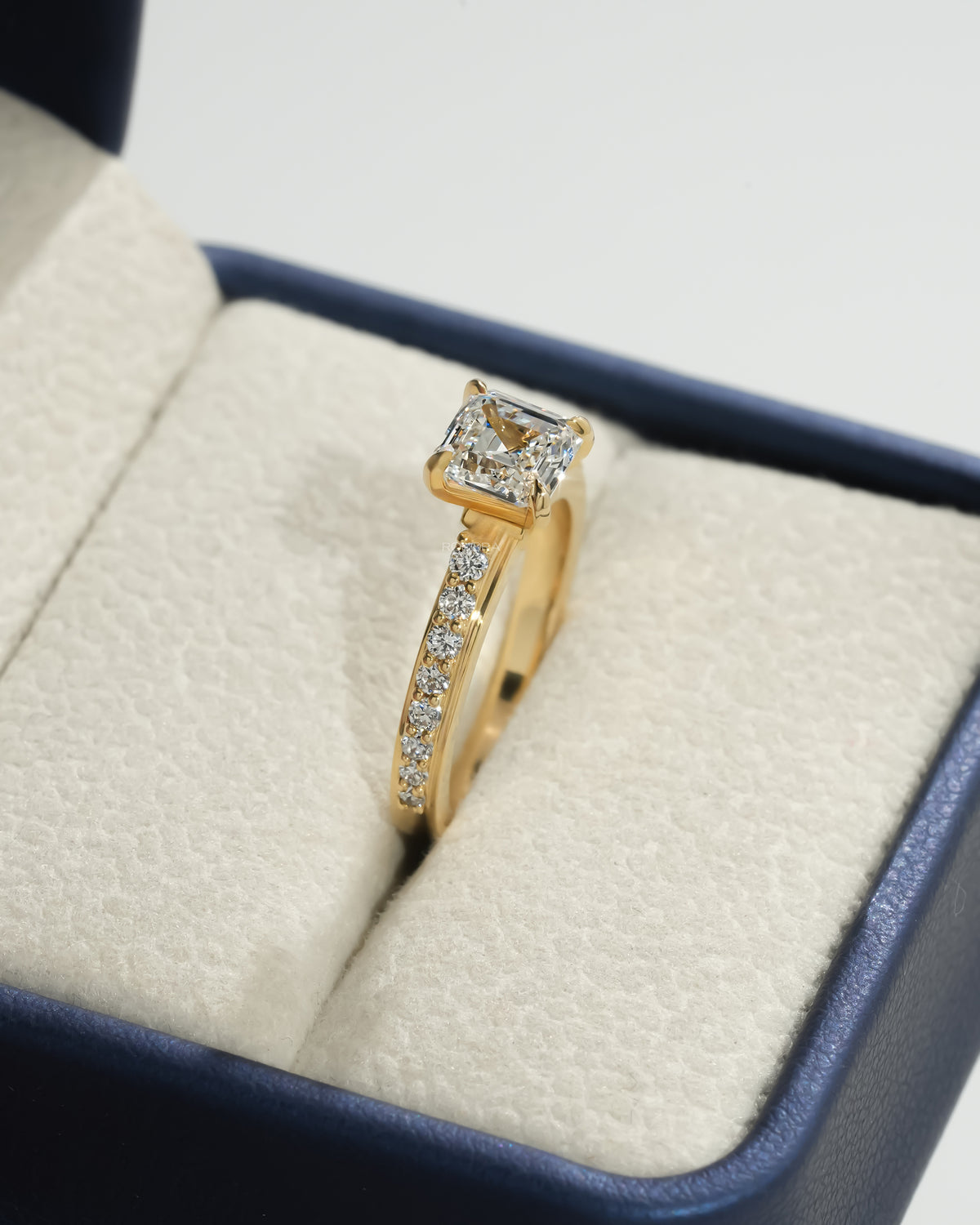 Side view of an asscher pave lab grown diamond ring in yellow gold, set in a cream and blue ring box