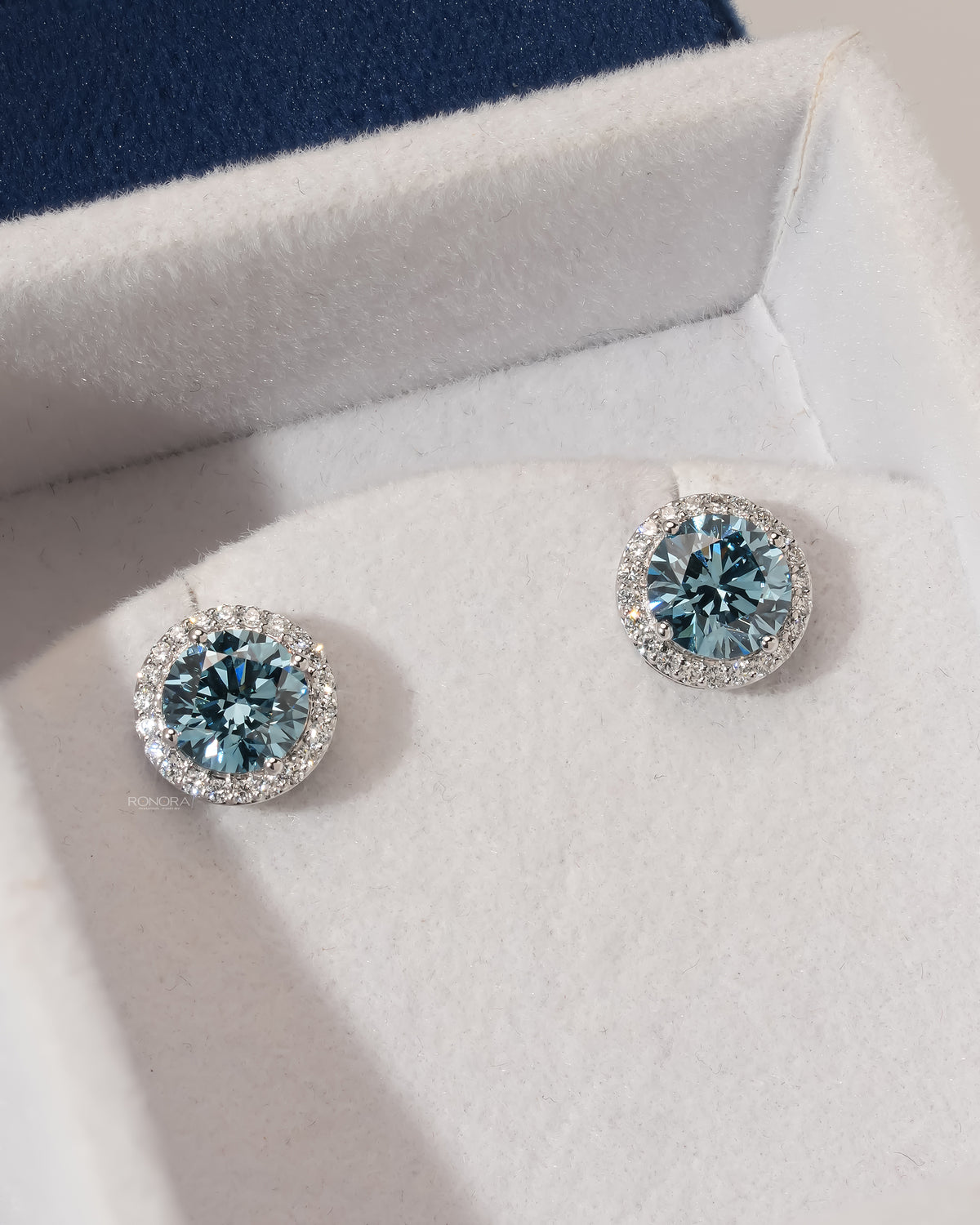 Pair of blue lab grown diamond halo stud earrings in white gold set in a white velvet jewelry box