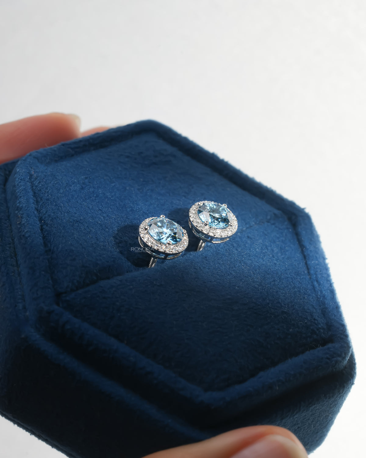 Blue lab grown diamond halo stud earrings in white gold resting in a navy velvet jewelry box held in hand