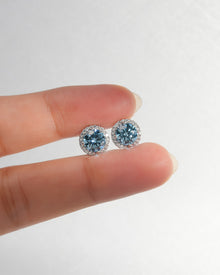 Blue lab grown diamond halo stud earrings in white gold held between two fingertips to show their size