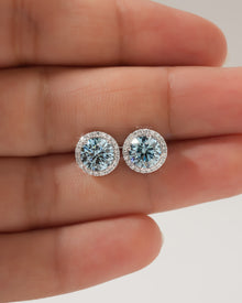 Close-up of a pair of blue lab grown diamond halo stud earrings in white gold resting on fingers