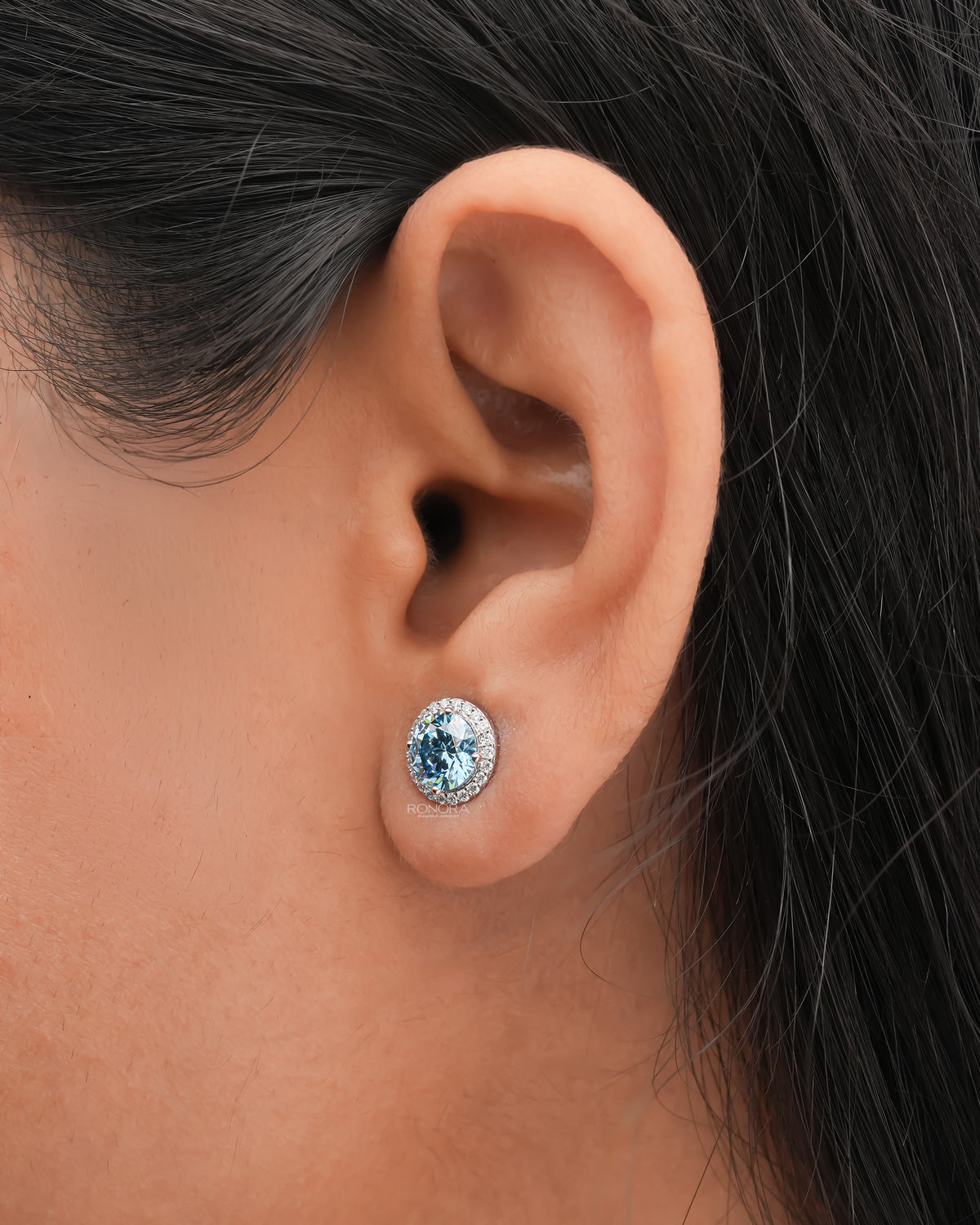 Close-up of a blue lab grown diamond halo stud earring worn on the ear, ringed by small sparkling diamonds