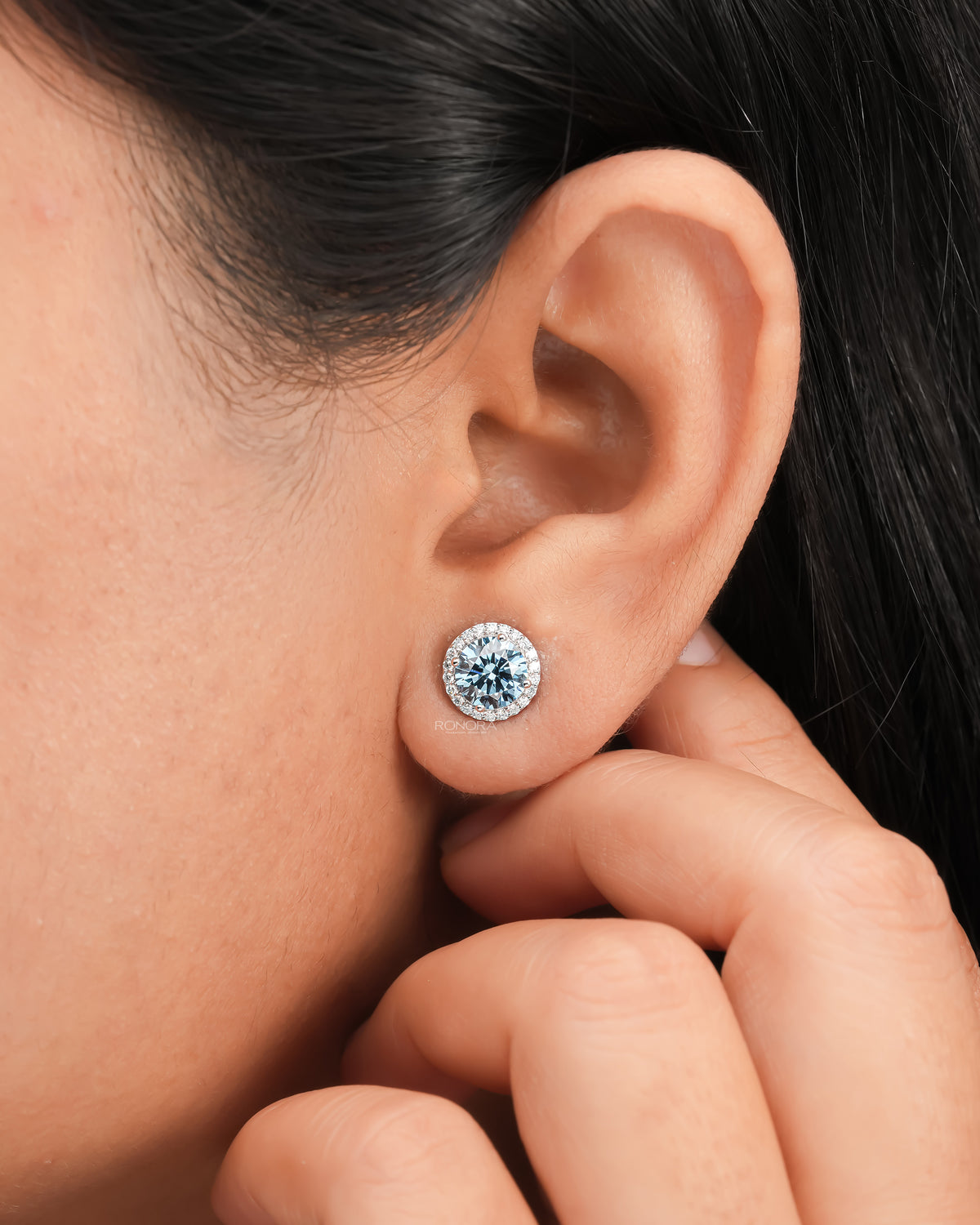 Blue lab grown diamond halo stud earring worn on the ear while a hand touches the lobe, white gold setting