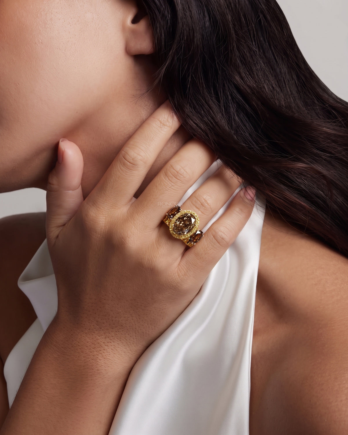 Woman wearing a brown oval lab grown diamond bridal set with a halo ring and yellow oval band, hand resting at her neck