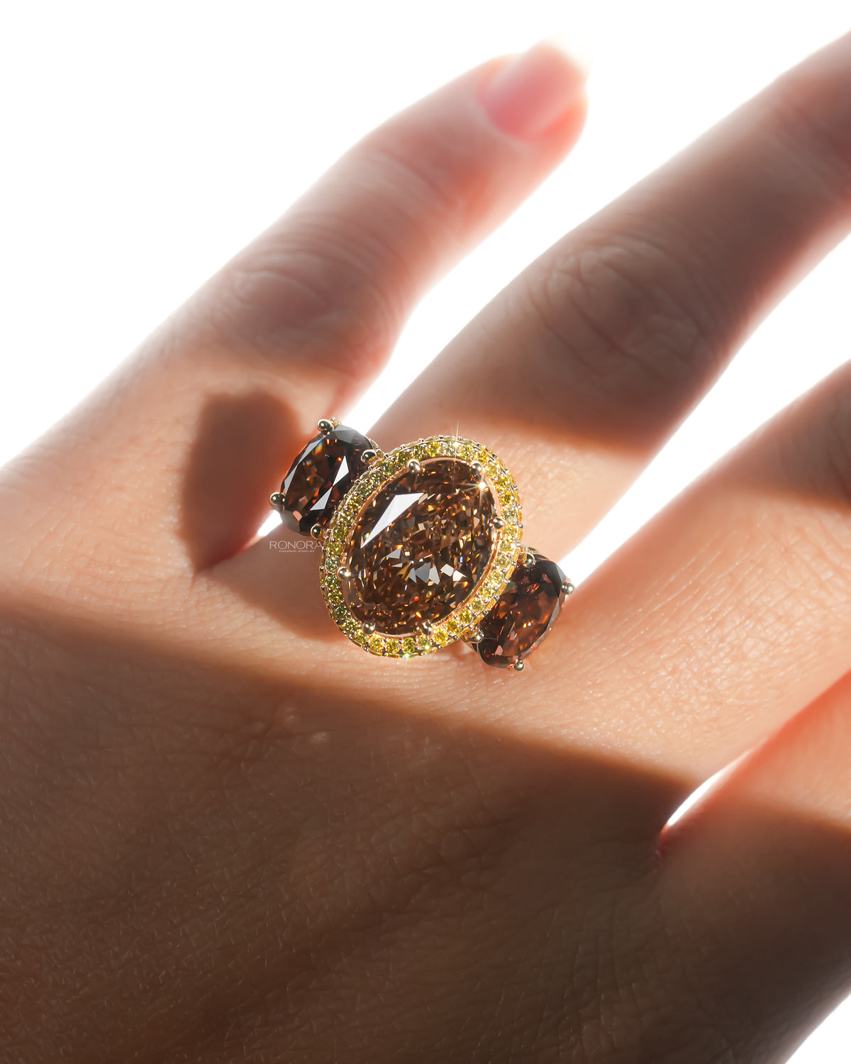 Sunlit hand wearing a brown oval lab grown diamond three stone ring, brown centre stone with yellow halo glowing