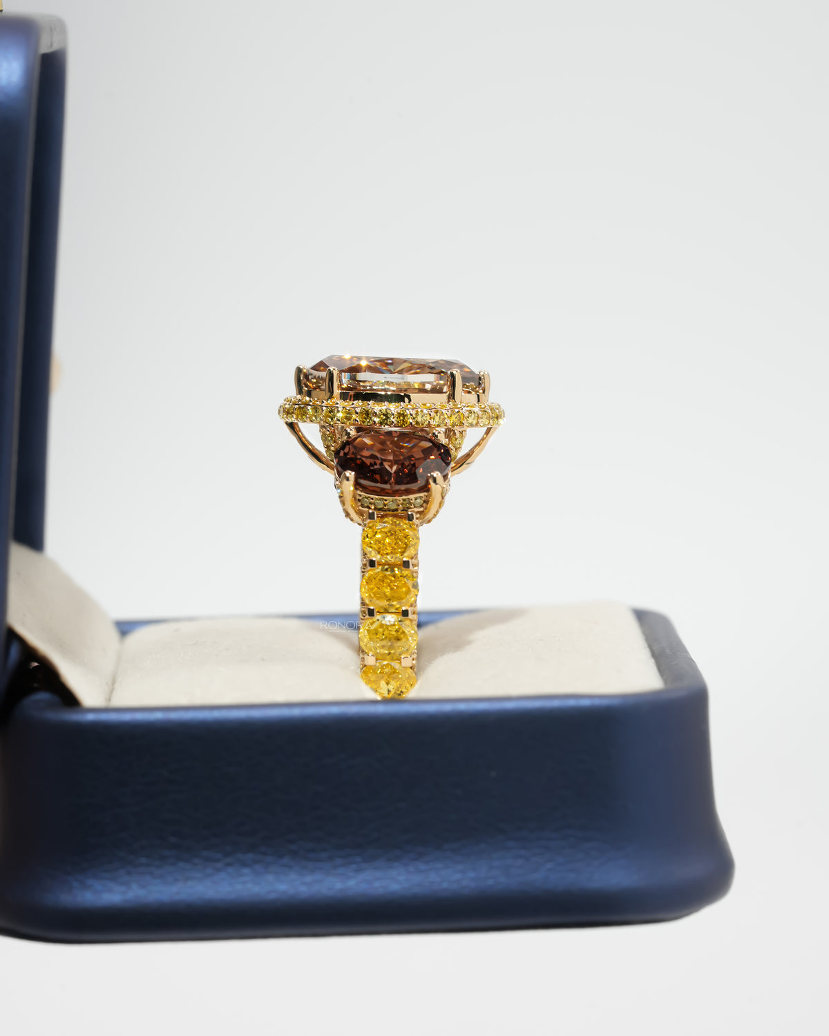 Side view of a brown oval lab grown diamond three stone ring with yellow halo, standing in a navy ring box